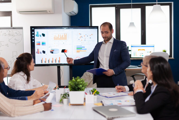 Successful team leader briefing pointing explaining project in conference room brainstorming. Corporate staff discussing new business application with colleagues looking at screen